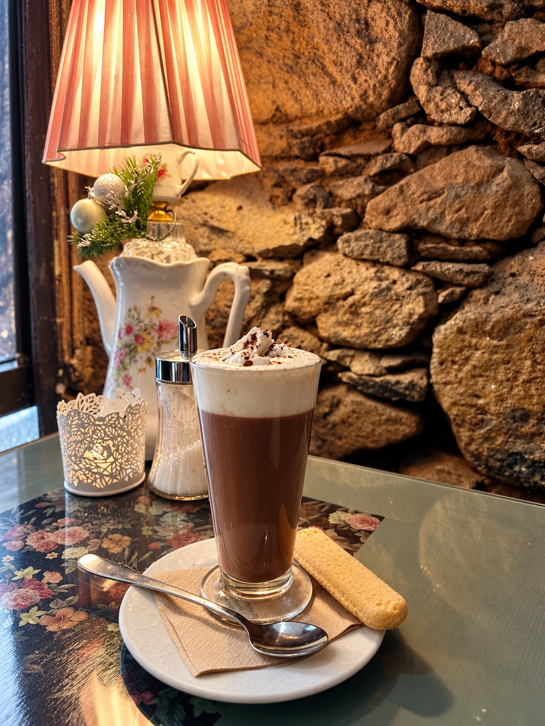 Chocolate con nata en vaso alto junto a una lámpara de pantalla rosa, una cafetera de porcelana y la pared de piedra