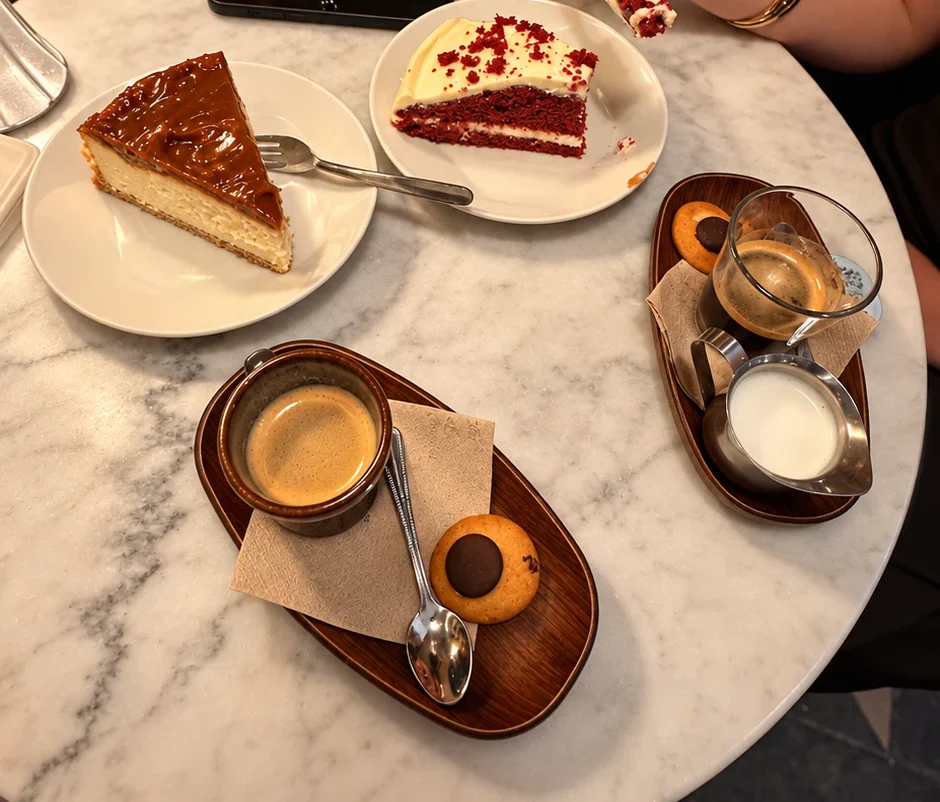 Mesa de mármol con una porción de tarta de queso con dulce de leche, otra de red velvet y dos cafés servidos en bandejitas de madera