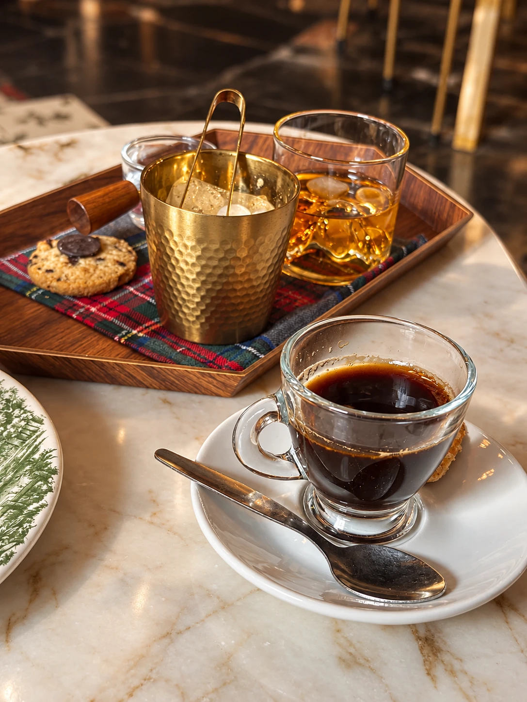 Un café solo en taza de cristal sobre una mesa de mármol, con una bandeja de madera detrás con whisky, cubitera dorada y una galleta